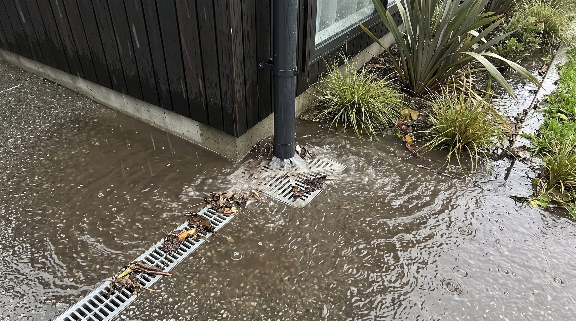 Downpipe draining into a debris-clogged channel drain during heavy rain